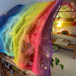 May include: A rainbow-coloured sheer fabric draped over a wooden structure, creating a play tent. The fabric is translucent, allowing light to shine through, and features a gradient of colours from blue to purple. Fairy lights are strung along the fabric, adding a whimsical touch.
