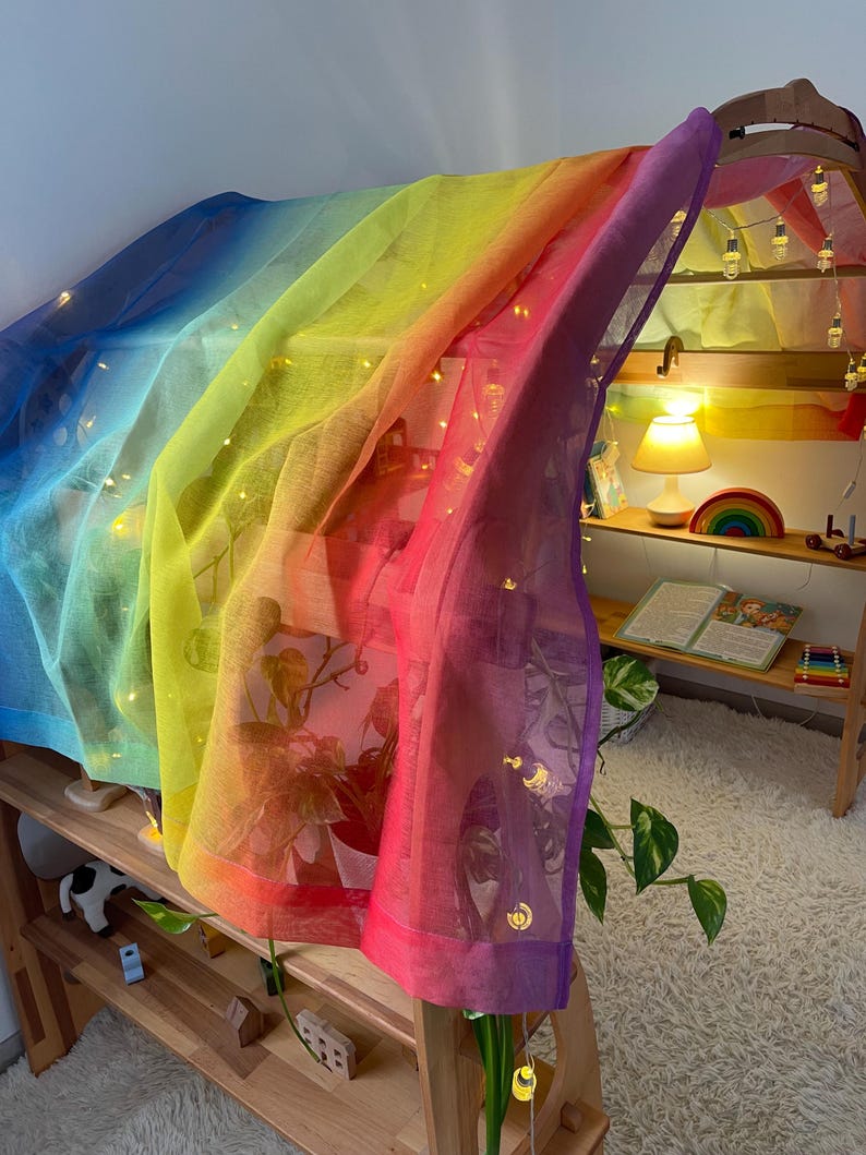 May include: A rainbow-colored sheer fabric draped over a wooden structure, creating a play tent. The fabric is translucent, allowing light to shine through, and features a gradient of colors from blue to purple. String lights are strung along the fabric, adding a whimsical touch.