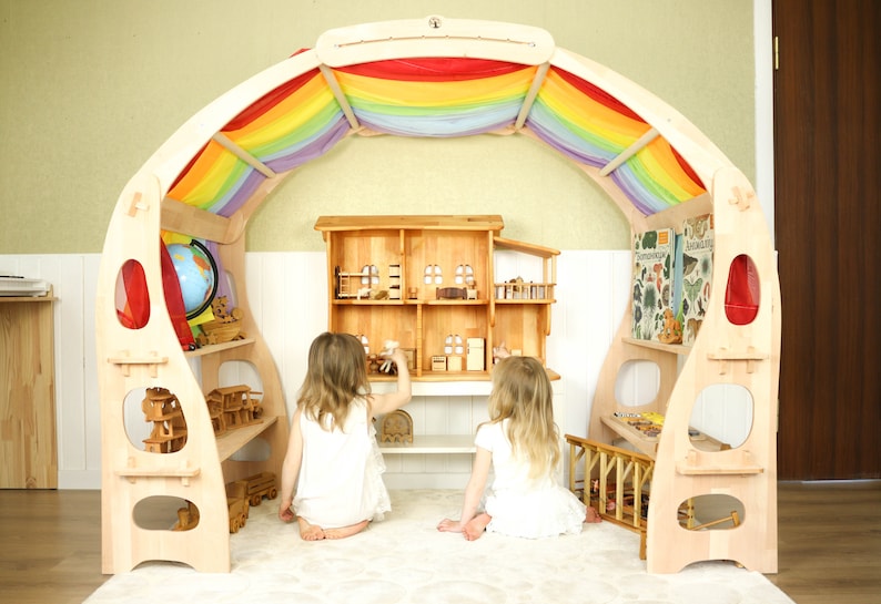 May include: Two children play in a wooden playhouse with a rainbow canopy. The playhouse has shelves and a dollhouse inside. The playhouse is made of natural wood and has a light wood finish.
