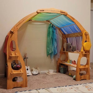 May include: A wooden play structure with a curved roof and shelves. The roof is covered with a blue and green fabric canopy. The structure has a small guitar, a basket of toys, and a few other items on the shelves.