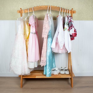 May include: A wooden clothing rack with a curved top and a shelf. Seven dresses and two shirts hang on the rack. The dresses are white, yellow, pink, and blue. The shirts are white and red. The rack is on a wooden floor.