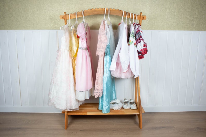 May include: A wooden clothing rack with a curved top and a shelf. Seven dresses and two shirts hang on the rack. The dresses are white, yellow, pink, and blue. The shirts are white and red. The rack is on a wooden floor.