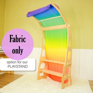 May include: Rainbow fabric play curtain with a wooden frame and shelves. The fabric is a gradient of colors from purple to red. The text "Fabric only" and "option for our PLAYSTAND" is printed on a pink circle.