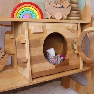 May include: A wooden toy washing machine with a round door and a small, open compartment. The door is open and there are some fabric items inside. The washing machine is part of a larger wooden playset.