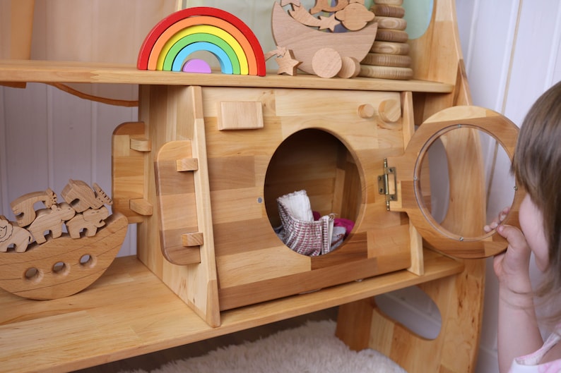 May include: A wooden toy washing machine with a round door and a small, open compartment. The door is open and there are some fabric items inside. The washing machine is part of a larger wooden playset.