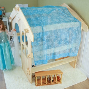 May include: A wooden playhouse with a blue and white fabric canopy. The playhouse has a small window and a bench inside. A child is standing in front of the playhouse.