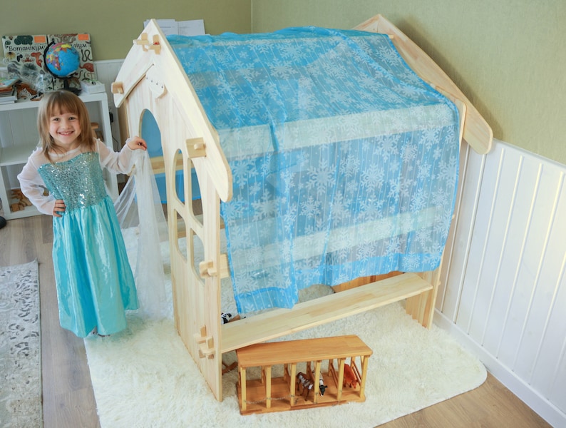 May include: A wooden playhouse with a blue and white fabric canopy. The playhouse has a small window and a bench inside. A child is standing in front of the playhouse.