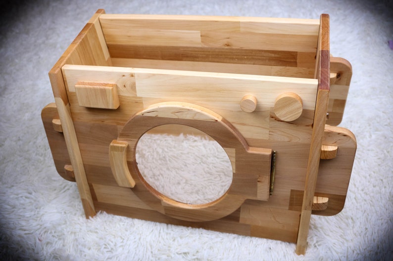 May include: A wooden toy washing machine with a round opening and two knobs. The toy is made of natural wood and has a rustic look.