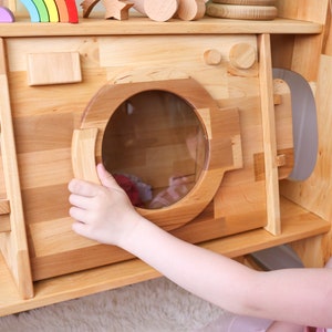 May include: A child's wooden play kitchen with a pretend washing machine. The washing machine has a clear plastic door and a wooden handle. The child is opening the door of the washing machine.