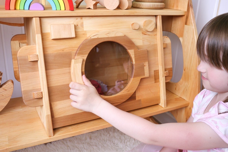 May include: A child's wooden play kitchen with a pretend washing machine. The washing machine has a clear plastic door and a wooden handle. The child is opening the door of the washing machine.