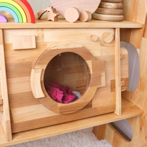May include: A wooden toy washing machine with a clear plastic door and a pink fabric item inside. The toy is part of a larger wooden playset with shelves.