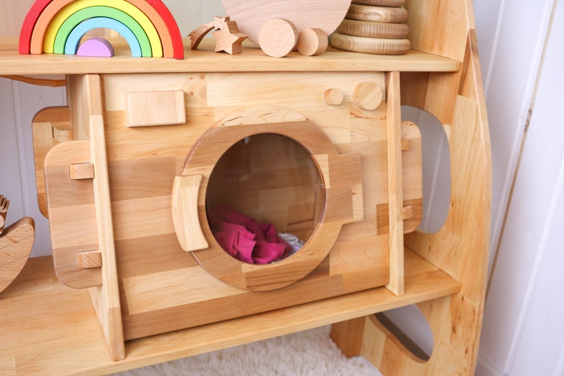 May include: A wooden toy washing machine with a clear plastic door and a pink fabric item inside. The toy is part of a larger wooden playset with shelves.