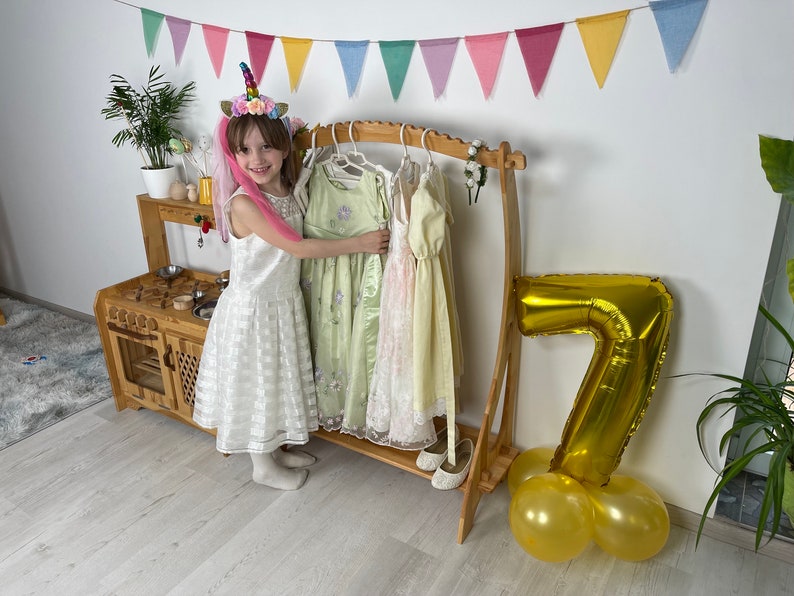 K&ouml;nnte beinhalten: Ein junges M&auml;dchen in einem wei&szlig;en Kleid und einem Einhorn-Stirnband steht vor einem Kleiderst&auml;nder aus Holz. Der St&auml;nder ist mit Kleidern gef&uuml;llt und es gibt einen goldenen Ballon mit der Nummer 7 und goldene Ballons auf dem Boden.