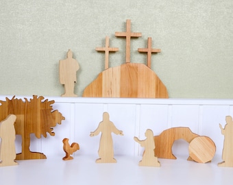 May include: Wooden figurines depicting the Easter story. The figures include Jesus, a rooster, a tree, a tomb, and angels. The figures are arranged on a white surface with a green background.