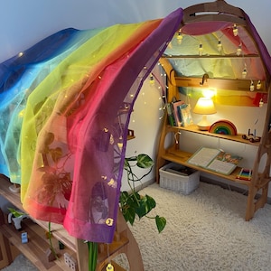 May include: A wooden play structure with a rainbow-colored fabric draped over it. The structure has shelves and a small lamp. There are string lights strung around the structure.