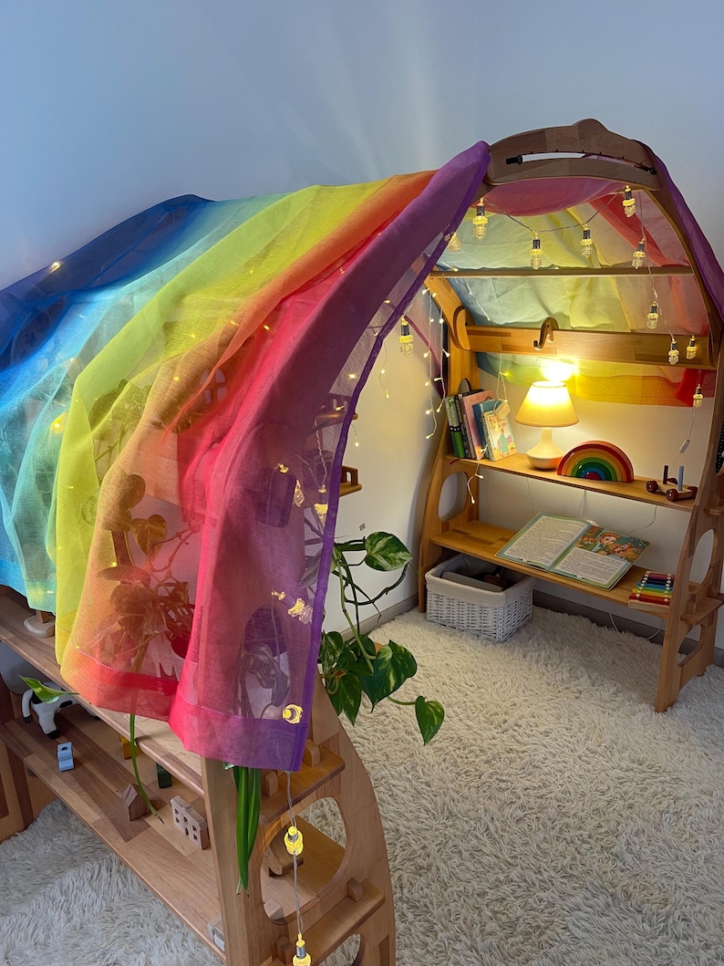 May include: A wooden play structure with a rainbow-colored fabric draped over it. The structure has shelves and a small lamp. There are string lights strung around the structure.