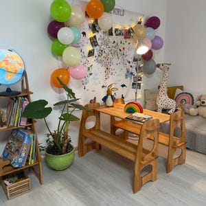 May include: A wooden children's table and bench set with two chairs. The table has two shelves and the bench has a back. The set is made of natural wood and has a light brown finish. There are balloons in the background.