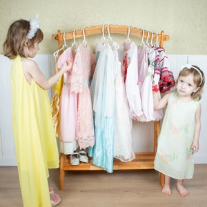 May include: Two young girls stand in front of a wooden clothing rack with a variety of colorful dresses hanging on hangers. The girl on the left is wearing a yellow dress and holding a pink dress. The girl on the right is wearing a light green dress with a floral pattern.