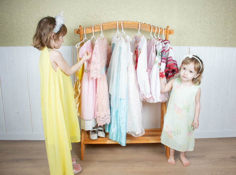 May include: Two young girls stand in front of a wooden clothing rack with a variety of colorful dresses hanging on hangers. The girl on the left is wearing a yellow dress and holding a pink dress. The girl on the right is wearing a light green dress with a floral pattern.