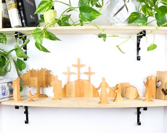 May include: Wooden figurines depicting the Easter story, including a cross, a tree, and figures of people. The figurines are arranged on a wooden shelf with a potted plant hanging over the shelf.