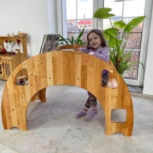 May include: A wooden archway play structure with a child sitting on the top. The archway has several openings and is made of natural wood. The child is wearing a purple shirt and is holding a pencil.