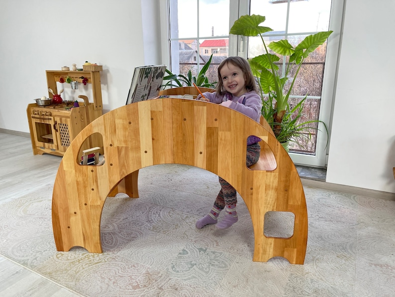 May include: A wooden archway play structure with a child sitting on the top. The archway has several openings and is made of natural wood. The child is wearing a purple shirt and is holding a pencil.