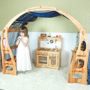 May include: A wooden playhouse with a blue fabric canopy and a play kitchen inside. The playhouse has shelves and a bench. A young girl is standing in front of the playhouse.