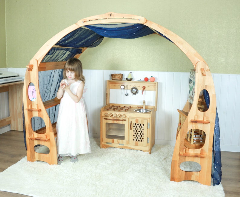 May include: A wooden playhouse with a blue fabric canopy and a play kitchen inside. The playhouse has shelves and a bench. A young girl is standing in front of the playhouse.