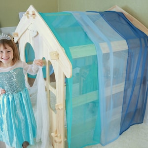 May include: A young girl dressed in a light blue princess costume stands in front of a wooden playhouse with blue and turquoise curtains. The playhouse has a small window and a door.