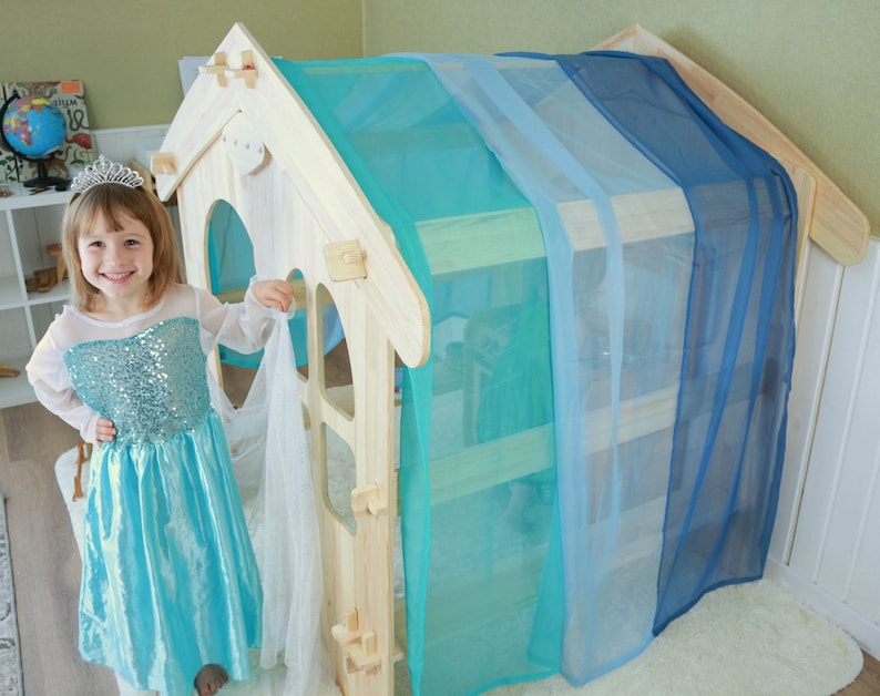 May include: A young girl dressed in a light blue princess costume stands in front of a wooden playhouse with blue and turquoise curtains. The playhouse has a small window and a door.