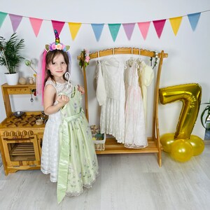 K&ouml;nnte beinhalten: Ein junges M&auml;dchen in einem wei&szlig;en Kleid mit gr&uuml;nem &Uuml;berzug und einem Einhorn-Stirnband steht vor einer Holzk&uuml;che und einem Kleiderst&auml;nder mit mehreren Kleidern, die daran h&auml;ngen. Ein goldener Ballon mit der Nummer 7 liegt auf dem Boden neben dem St&auml;nder.