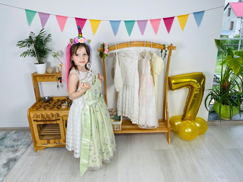 K&ouml;nnte beinhalten: Ein junges M&auml;dchen in einem wei&szlig;en Kleid mit gr&uuml;nem &Uuml;berzug und einem Einhorn-Stirnband steht vor einer Holzk&uuml;che und einem Kleiderst&auml;nder mit mehreren Kleidern, die daran h&auml;ngen. Ein goldener Ballon mit der Nummer 7 liegt auf dem Boden neben dem St&auml;nder.