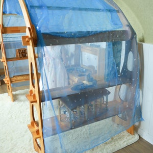 May include: A wooden play structure with shelves and a blue sheer curtain with gold stars. The structure is designed for children's play and is made of natural wood.