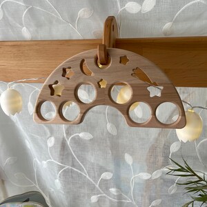 May include: A wooden rainbow-shaped toy with cutouts of stars, moons, and circles. The toy is hanging from a wooden hook.