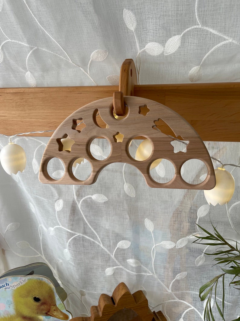 May include: A wooden rainbow-shaped toy with cutouts of stars, moons, and circles. The toy is hanging from a wooden hook.