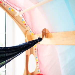 May include: A wooden play structure with a blue and gold fabric hanging from a hook. The fabric has a star pattern. The structure is decorated with colorful pom-poms.