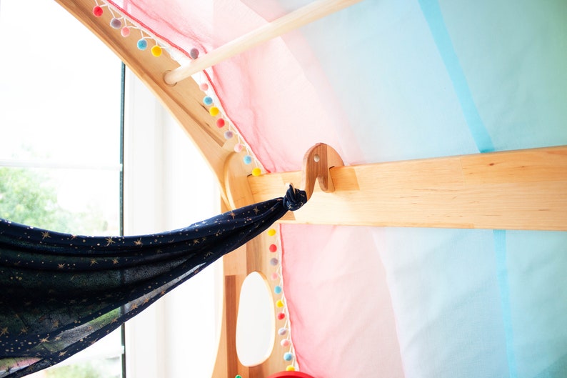 May include: A wooden play structure with a blue and gold fabric hanging from a hook. The fabric has a star pattern. The structure is decorated with colorful pom-poms.