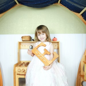 May include: A young girl in a white dress holds a wooden letter E with the name "Emma" carved into it. The letter is in a playroom setting with wooden shelves and toys.