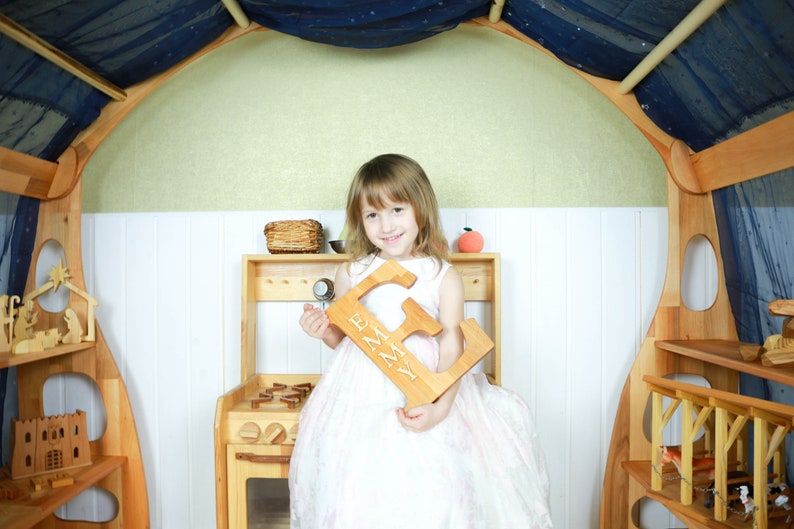 May include: A young girl in a white dress holds a wooden letter E with the name "Emma" carved into it. The letter is in a playroom setting with wooden shelves and toys.