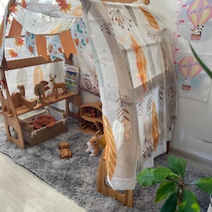 May include: A wooden play tent with a white and orange patterned fabric canopy. The tent is decorated with a garland of orange and white fabric flags. The tent is set up in a room with a gray rug and a wooden floor.