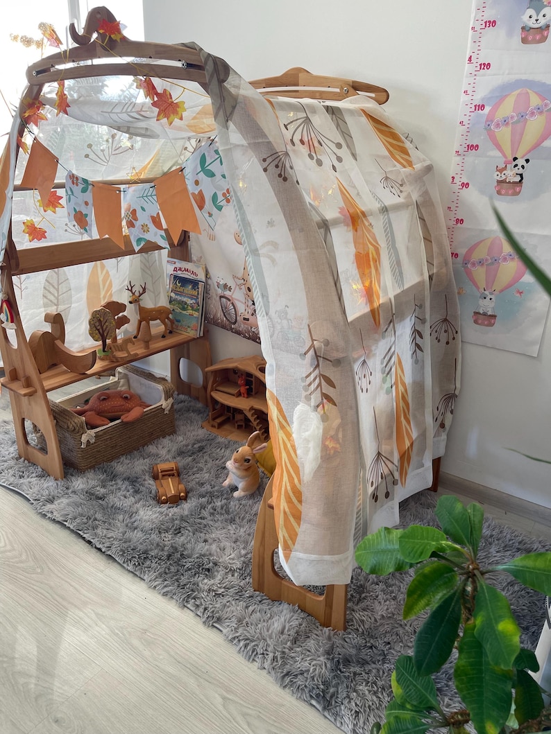 May include: A wooden play tent with a white and orange patterned fabric canopy. The tent is decorated with a garland of orange and white fabric flags. The tent is set up in a room with a gray rug and a wooden floor.