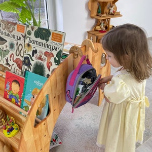 May include: A wooden children's play structure with a bookshelf, a small wooden dollhouse, and a purple backpack hanging on a hook. A young girl in a yellow dress is standing next to the structure.