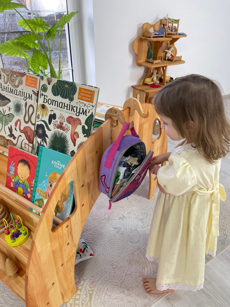 May include: A wooden children's play structure with a bookshelf, a small wooden dollhouse, and a purple backpack hanging on a hook. A young girl in a yellow dress is standing next to the structure.