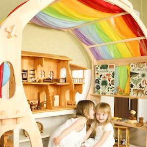 May include: Two children play in a wooden playhouse with a rainbow fabric canopy. The playhouse has shelves with books titled "Botanicum" and "Animalum".