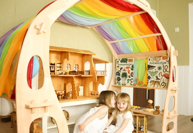 May include: Two children play in a wooden playhouse with a rainbow fabric canopy. The playhouse has shelves with books titled "Botanicum" and "Animalum".