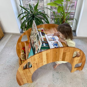 May include: A wooden children's table with a curved design and a built-in storage shelf. The table has a light wood finish and is perfect for a child's playroom or bedroom. A young child is sitting at the table, reading a book and using colored pencils.