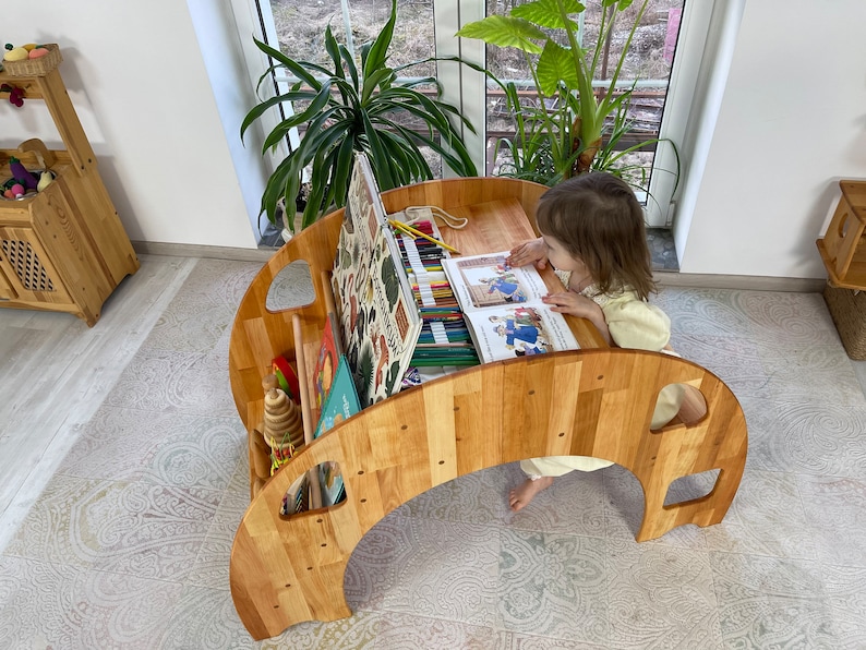 May include: A wooden children's table with a curved design and a built-in storage shelf. The table has a light wood finish and is perfect for a child's playroom or bedroom. A young child is sitting at the table, reading a book and using colored pencils.