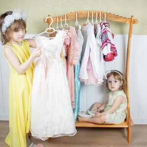 K&ouml;nnte beinhalten: Zwei junge M&auml;dchen spielen Verkleiden. Ein M&auml;dchen tr&auml;gt ein gelbes Kleid und h&auml;lt ein wei&szlig;es Spitzenkleid. Das andere M&auml;dchen sitzt auf einem Holz-Kleiderst&auml;nder und tr&auml;gt ein gr&uuml;nes Kleid. Der St&auml;nder ist mit verschiedenen Kleidern und Hemden gef&uuml;llt.