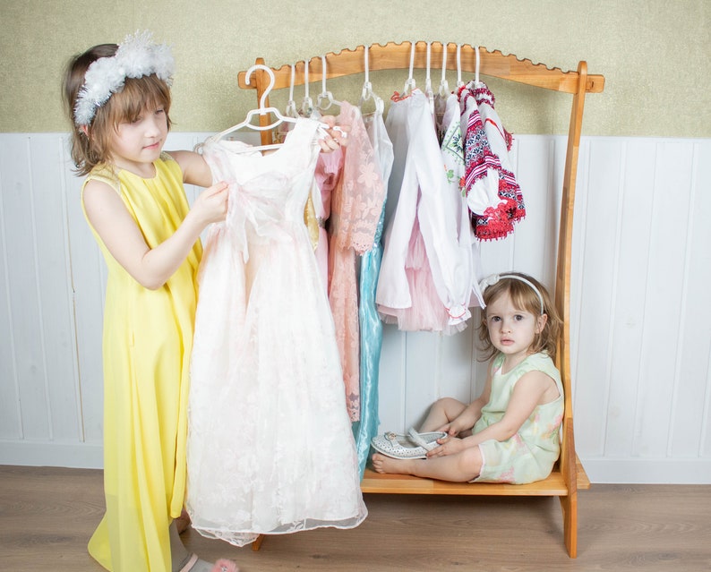 K&ouml;nnte beinhalten: Zwei junge M&auml;dchen spielen Verkleiden. Ein M&auml;dchen tr&auml;gt ein gelbes Kleid und h&auml;lt ein wei&szlig;es Spitzenkleid. Das andere M&auml;dchen sitzt auf einem Holz-Kleiderst&auml;nder und tr&auml;gt ein gr&uuml;nes Kleid. Der St&auml;nder ist mit verschiedenen Kleidern und Hemden gef&uuml;llt.