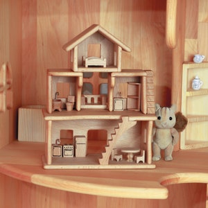 May include: A wooden dollhouse with three stories, featuring miniature furniture and a staircase. The dollhouse is made of light-colored wood and includes a small, plush animal figure. The overall design is simple and charming.